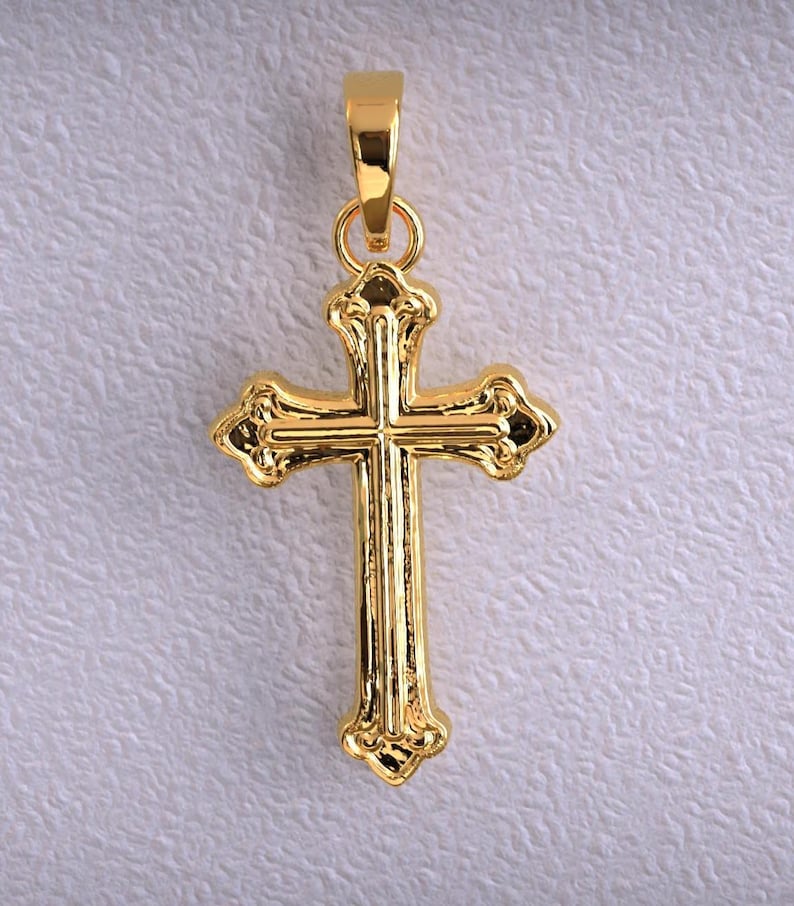Puede incluir: Un colgante dorado con forma de cruz. La cruz tiene detalles ornamentados y un lazo en la parte superior para una cadena. El colgante se muestra sobre un fondo texturizado blanquecino.