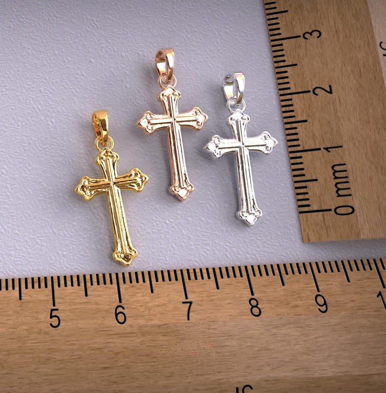 Puede incluir: Tres colgantes decorativos de cruz en oro, oro rosa y plata. Cada cruz tiene un dise&ntilde;o detallado y un lazo para una cadena. Los colgantes se muestran sobre una superficie gris junto a una regla de madera.