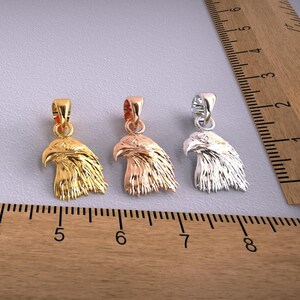 May include: Three metal eagle head pendants in gold, rose gold, and silver. Each pendant features a detailed carving of an eagle's head with a loop for a chain. The pendants are displayed on a gray surface next to a wooden ruler.