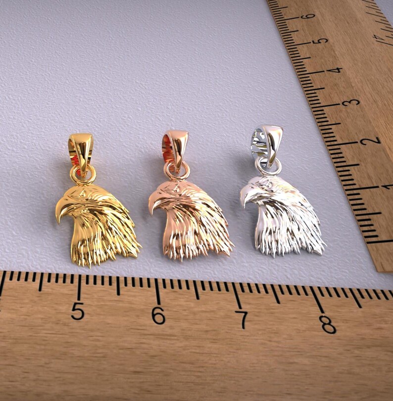 May include: Three metal eagle head pendants in gold, rose gold, and silver. Each pendant features a detailed carving of an eagle's head with a loop for a chain. The pendants are displayed on a gray surface next to a wooden ruler.
