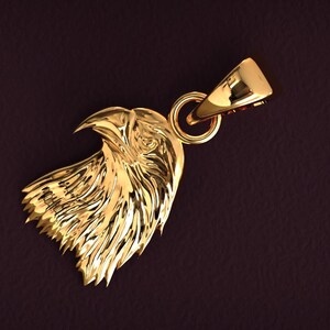 May include: A gold-colored pendant in the shape of an eagle's head. The pendant features detailed feather and beak carvings, with a loop for a chain. The background is a dark, textured brown.