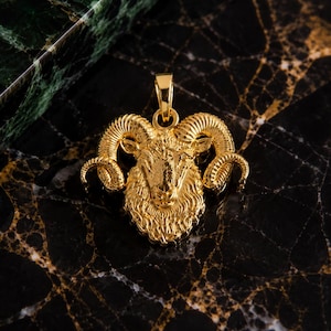 May include: A gold-toned metal pendant shaped like a ram head with detailed curled horns and textured fur. The pendant features a small circular bail at the top for attachment to a chain, showcasing a polished, reflective surface throughout the design.
