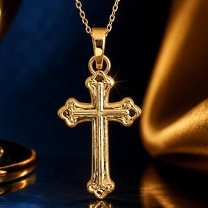 Puede incluir: Un collar con colgante dorado con un dise&ntilde;o de cruz detallado. La cruz tiene detalles ornamentados y cuelga de una delicada cadena. La joya se exhibe sobre un fondo azul oscuro y dorado, resaltando su brillo.