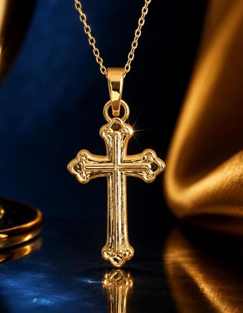 Puede incluir: Un collar con colgante dorado con un dise&ntilde;o de cruz detallado. La cruz tiene detalles ornamentados y cuelga de una delicada cadena. La joya se exhibe sobre un fondo azul oscuro y dorado, resaltando su brillo.