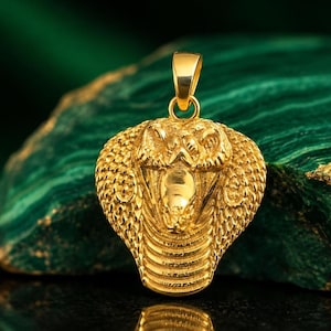 May include: A gold-toned pendant shaped like a cobra with its hood flared. The surface features detailed scales and a smooth, raised head section. A small, circular bail is attached to the top of the snake's head for hanging on a chain or cord.