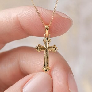 Puede incluir: Un collar con colgante de cruz de color dorado. La cruz tiene un dise&ntilde;o ornamentado con detalles decorativos. La cadena del collar es delicada y tambi&eacute;n de color dorado. El colgante est&aacute; sostenido por los dedos de una persona.
