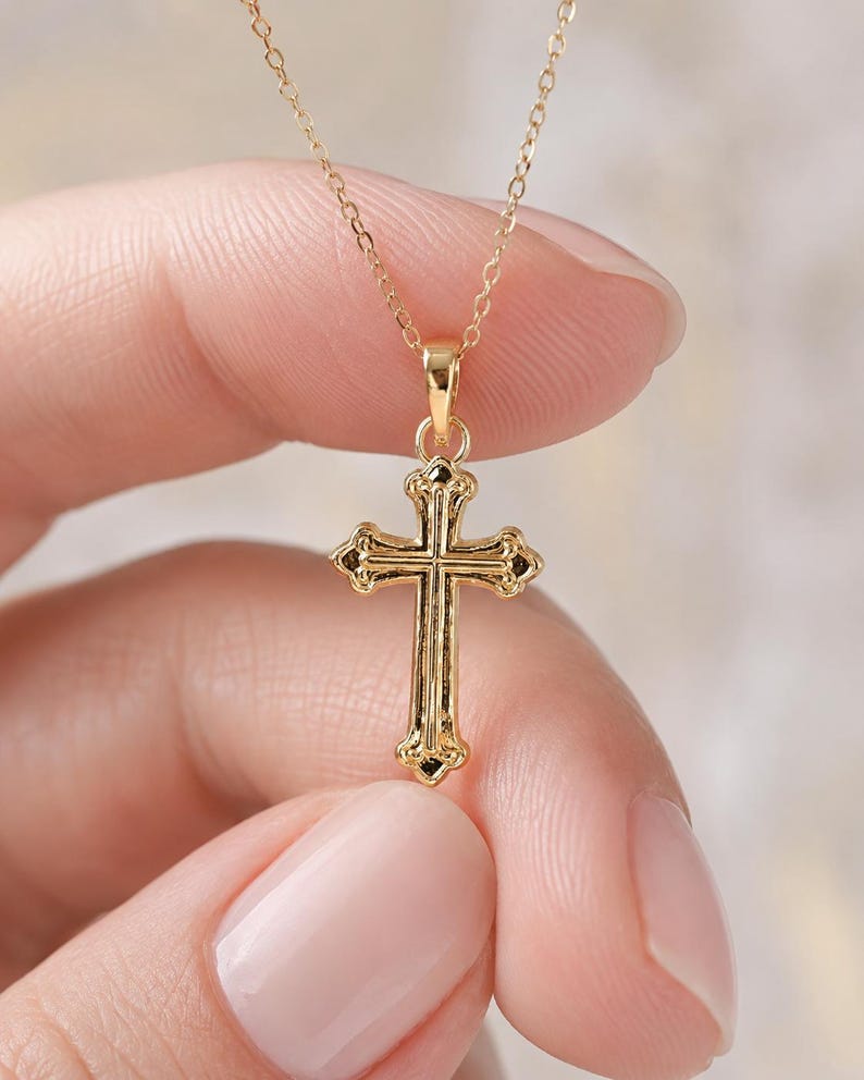 Puede incluir: Un collar con colgante de cruz de color dorado. La cruz tiene un dise&ntilde;o ornamentado con detalles decorativos. La cadena del collar es delicada y tambi&eacute;n de color dorado. El colgante est&aacute; sostenido por los dedos de una persona.