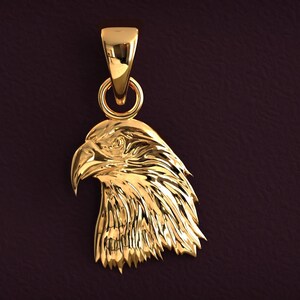 May include: A gold-colored pendant in the shape of an eagle's head. The pendant has a detailed design, with textured feathers and a sharp beak. It includes a loop for a chain and a bail for hanging.