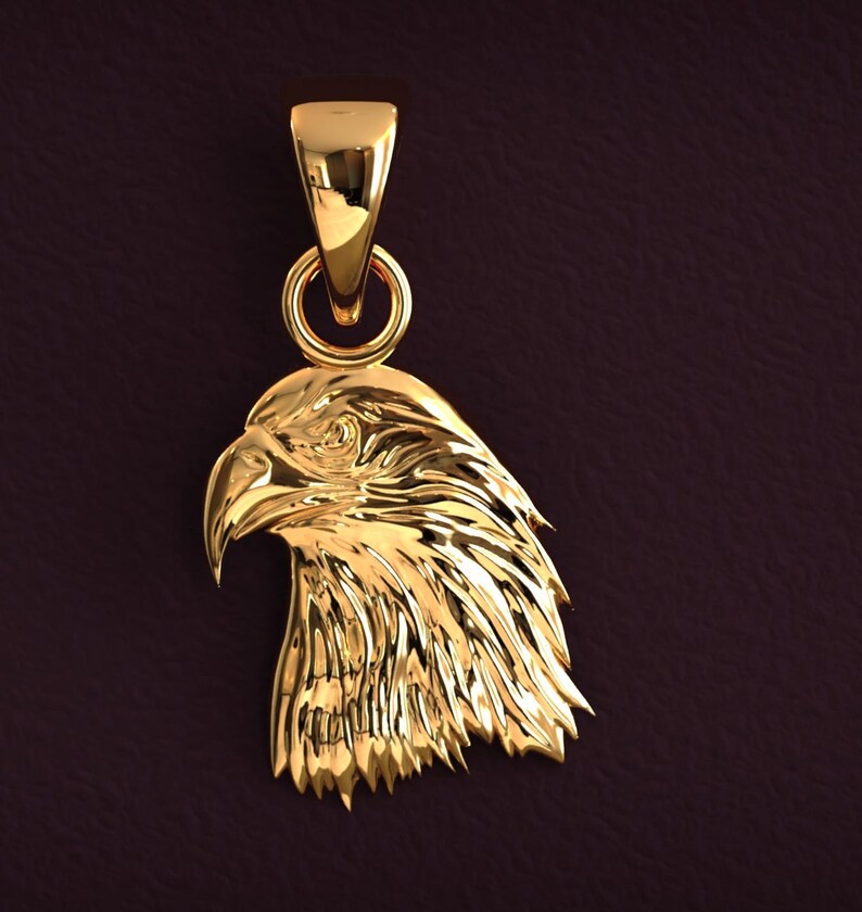 May include: A gold-colored pendant in the shape of an eagle's head. The pendant has a detailed design, with textured feathers and a sharp beak. It includes a loop for a chain and a bail for hanging.
