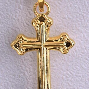 Puede incluir: Colgante dorado en forma de cruz. La cruz tiene detalles ornamentados y un lazo para una cadena. El colgante es probablemente de metal y tiene un acabado brillante.