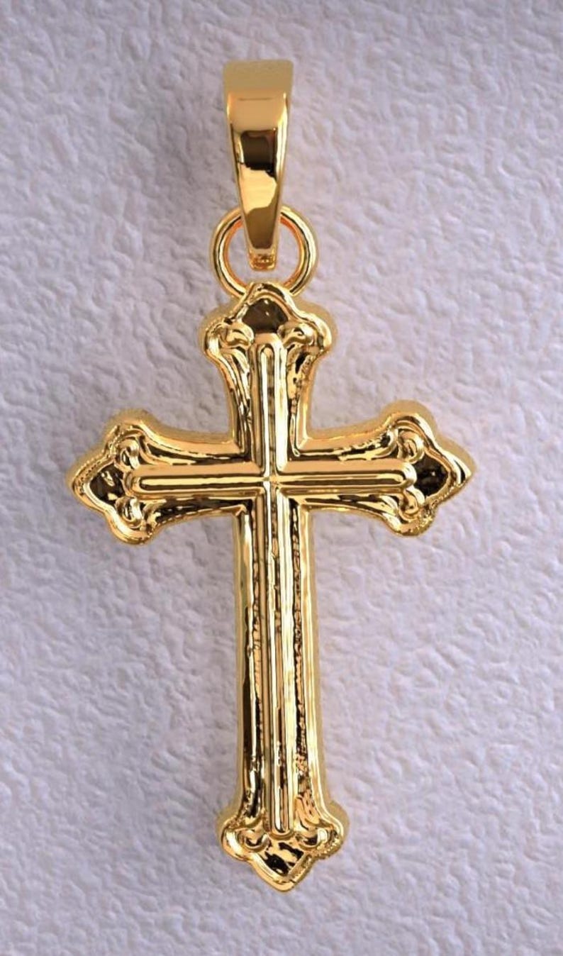 Puede incluir: Colgante dorado en forma de cruz. La cruz tiene detalles ornamentados y un lazo para una cadena. El colgante es probablemente de metal y tiene un acabado brillante.
