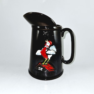 May include: Black ceramic pitcher with a rooster painted on the side. The rooster is red, white, and blue and is standing on a red roof.