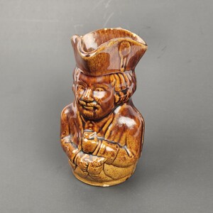 May include: A brown ceramic pitcher shaped like a person wearing a hat. The pitcher has a detailed face and is holding a small object in its hands.