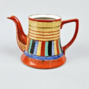 May include: A red ceramic teapot with a wide spout and a handle. The teapot is decorated with a colorful geometric pattern in shades of blue, green, yellow, and black.