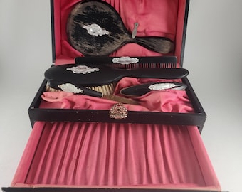 May include: A black wooden vanity set with a drawer and a hinged lid. The set includes a hand mirror, a hairbrush, a comb, and a nail file. The interior of the set is lined with pink fabric.
