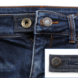 May include: A close-up of a pair of blue jeans with a metal button and a decorative button. The decorative button is a circle with a star design and a laurel wreath around it. The button is attached to a piece of denim fabric.