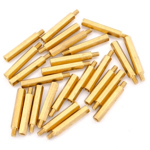 May include: A group of 20 gold-coloured metal standoffs, also known as spacers, are scattered on a white surface. The standoffs are hexagonal in shape and have a threaded hole on one end.