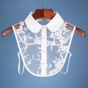 May include: A white lace collar with a Peter Pan collar design. The collar features a delicate floral lace overlay and a button-down front. The collar is displayed on a wooden hanger against a blue background.