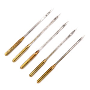 以下が含まれることがあります： 銀色の先端を持つ金色のミシン針が5本。針は一列に並んでおり、先端は右を向いています。針はすべて同じサイズで、側面に「SINGER 107RS」と印刷されています。