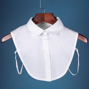 以下が含まれることがあります： ボタン留めのフロントと調節可能なストラップが付いた、取り外し可能な白い襟。襟は青い背景のウッドハンガーにかかっています。