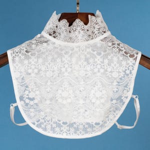 May include: White lace collar with a scalloped edge. The collar is on a hanger and has adjustable straps.