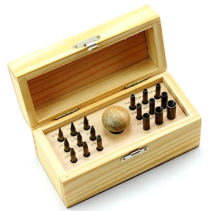 May include: A wooden box with a hinged lid, containing various metal punches and a wooden ball. The punches are of different sizes, labeled in mm, and are arranged in a wooden tray. The box is made of light-colored wood.