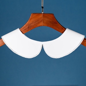 May include: White cotton peter pan collar with a scalloped edge. The collar is hanging on a wooden hanger.