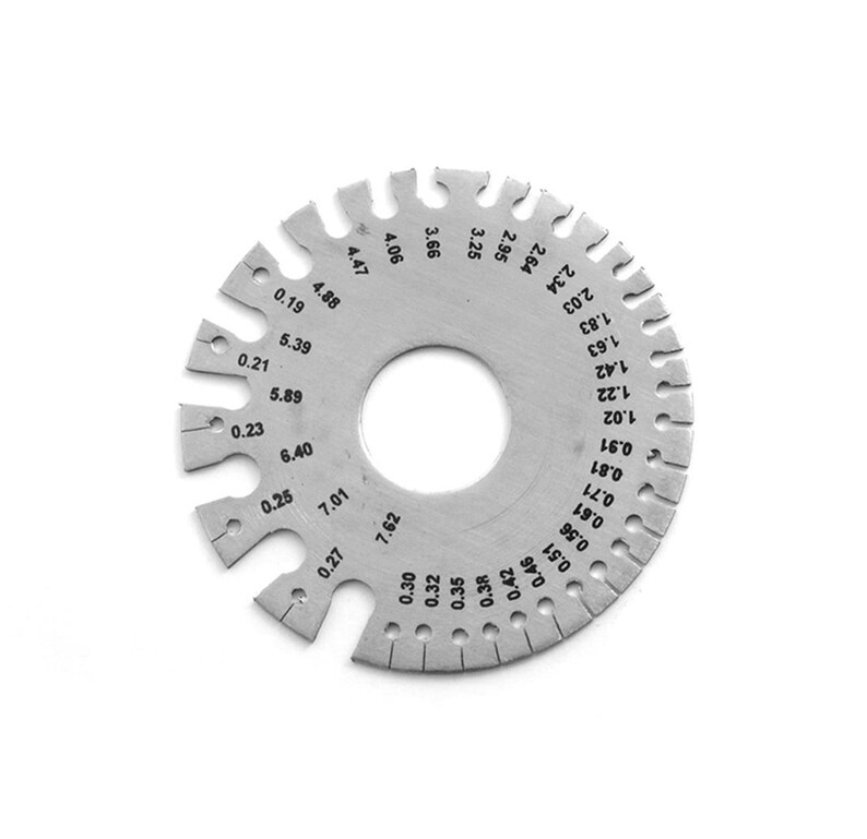 Puede incluir: Una galga de metal plateado con un agujero circular en el centro y una serie de marcas alrededor del borde. Las marcas incluyen n&uacute;meros y fracciones de pulgada, como 0,19, 0,21, 0,23, 0,25, 0,27, 0,30, 0,32, 0,35, 0,38, 0,42, 0,46, 0,51, 0,56, 0,61, 0,71, 0,81, 0,91, 1,02, 1,22, 1,42, 1,63, 1,83, 2,03, 2,34, 2,64, 2,95, 3,25, 3,66, 4,06, 4,47, 4,88, 5,39, 5,89, 6,40, 7,01 y 7,62.