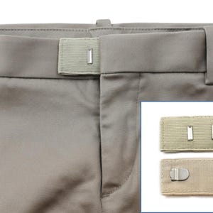 May include: Two beige fabric belt extenders with metal clasps. The extenders are shown on a pair of beige trousers.