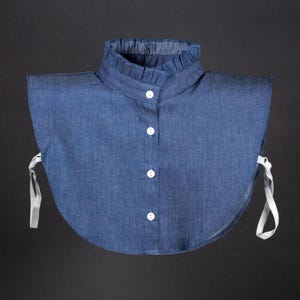 Puede incluir: Un cuello de mezclilla con un cuello de volantes y botones blancos. El cuello tiene lazos blancos a cada lado.