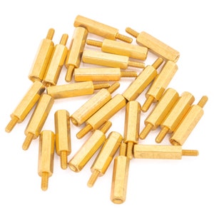 May include: A group of gold-coloured metal standoffs, also known as spacers, are scattered on a white surface. These standoffs are used to elevate components and create space between them.