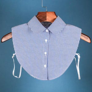 May include: A white and blue striped fabric collar with white buttons and white ties on each side. The collar is hanging on a wooden hanger.