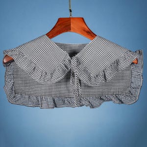 May include: A black and white gingham fabric collar with a ruffled edge. The collar has a tie at the center front.