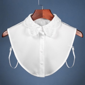 Könnte beinhalten: Abnehmbarer weißer Kragen mit Spitzenbesatz. Der Kragen hat eine abgerundete Form und ist zum Tragen über einem Hemd oder einer Bluse gedacht.