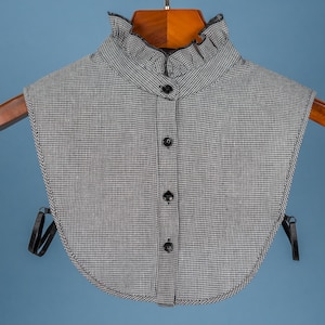 Könnte beinhalten: Ein schwarzer und weißer Gingham-Kragen aus Stoff mit einem Rüschenkragen und vier schwarzen Knöpfen an der Vorderseite.