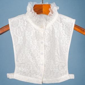 May include: A white lace collar with a frilled neckline and a pearl button fastening. The collar is buttoned down the front and features a delicate floral pattern. The collar is displayed on a wooden hanger against a blue backdrop.