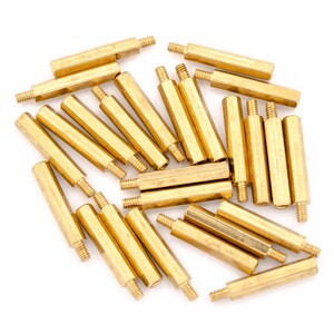 May include: A group of 20 gold-coloured metal standoffs, also known as spacers, are scattered on a white surface. The standoffs are cylindrical with a hexagonal head on one end and a threaded hole on the other end.