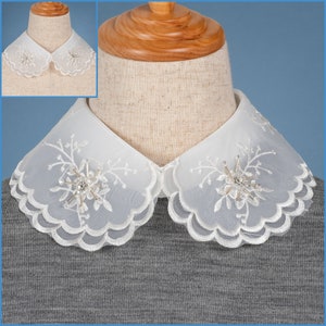 May include: White lace collar with scalloped edges and delicate floral embroidery. The collar features a small, sparkling rhinestone embellishment in the center.
