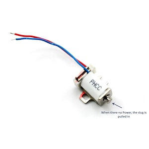 Può includere: Un piccolo componente elettrico bianco con fili rossi e blu che si estendono da esso. Il componente ha la scritta "PHCC" stampata sul lato. Un piccolo pezzo di metallo è attaccato al componente. È visibile anche il testo "When there is no Power, the slug is pulled in".