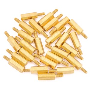 May include: A group of 20 gold-coloured metal standoffs, also known as spacers, are scattered on a white surface. The standoffs are hexagonal in shape and have a threaded hole on each end.
