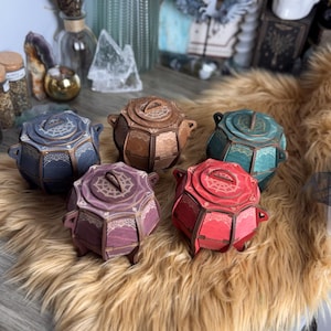May include: Five decorative wooden boxes in various colors: blue, brown, teal, purple, and red. Each box has a unique design on the lid and sides, with small feet. The boxes are arranged on a fluffy, light brown surface.