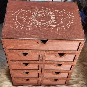 May include: A wooden storage box with eight small drawers. The top of the box features a laser-cut design of the sun, moon, and stars. The box is stained a reddish-brown color and sits on a fluffy surface.