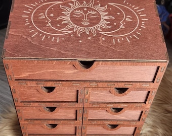 May include: A wooden storage box with eight small drawers. The top of the box features a laser-cut design of the sun, moon, and stars. The box is stained a reddish-brown color and sits on a fluffy surface.