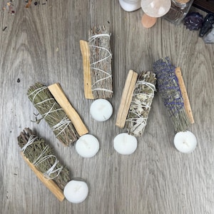 May include: An overhead shot of several bundles of sage, palo santo sticks, and small white candles arranged on a wooden surface. The sage bundles are tied with white string, and the palo santo sticks are light brown. The candles are round.