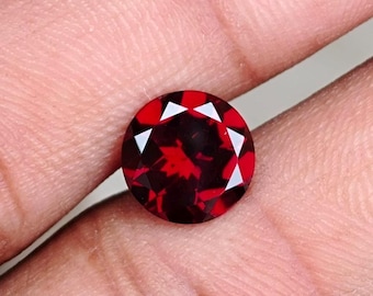 Puede incluir: Una gema redonda, facetada, de color rojo intenso. La gema tiene un corte brillante con facetas triangulares que reflejan la luz. La piedra se sostiene en una mano, mostrando su color y claridad. La gema parece ser un granate.