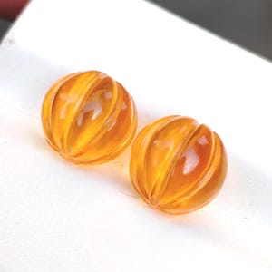 Può includere: Due perline rotonde color ambra con scanalature verticali, simili a mini zucche. Le perline traslucide lasciano passare la luce, su sfondo bianco. Queste perline potrebbero essere utilizzate per la creazione di gioielli.