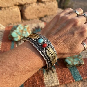 Peut inclure: Un bracelet manchette argenté orné d'une pierre turquoise ovale centrale et d'une pierre rouge ovale plus petite. La bande métallique présente des gravures dorées en forme de feuilles, des bords festonnés et de petites sphères argentées.