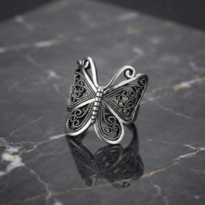 Peut inclure: Une bague en métal argenté en forme de papillon avec des découpes en filigrane complexes sur les ailes. La surface du métal présente une finition oxydée et sombre dans les zones en creux, soulignant les détails des volutes et des motifs.