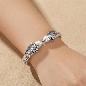 May include: A silver-toned cuff bracelet featuring two fish heads facing each other at the opening. The metal surface is textured with detailed scale patterns along the entire length of the band, creating a continuous design of two swimming fish.