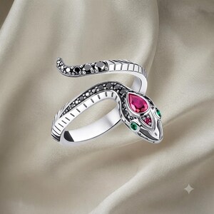Peut inclure: Une bague serpent argentée au design enveloppant ornée de pierres noires le long du corps. La tête du serpent est accentuée par une pierre rouge en forme de poire, une petite pierre rouge ronde et deux pierres vertes pour les yeux.